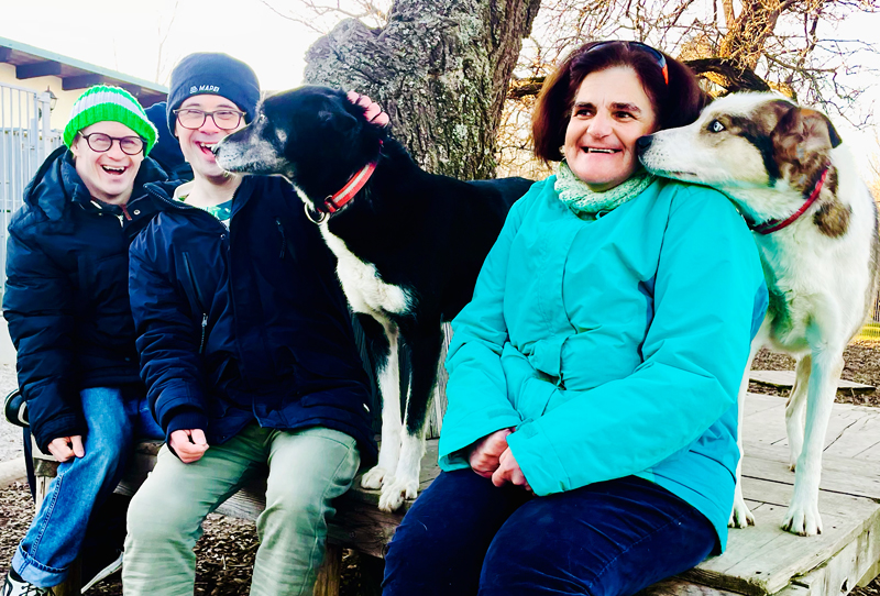 Eine Frau und zwei Männer mit besonderen Bedürfnissen freut sich über den Besuch bei den Huskys. Sie sitzen auf einer Bank und werden von den Huskys liebkost.
